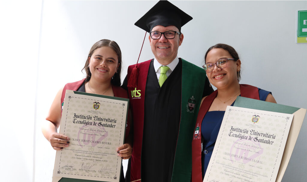 Dos graduadas muestran sus diplomas junto a un directivo con toga.