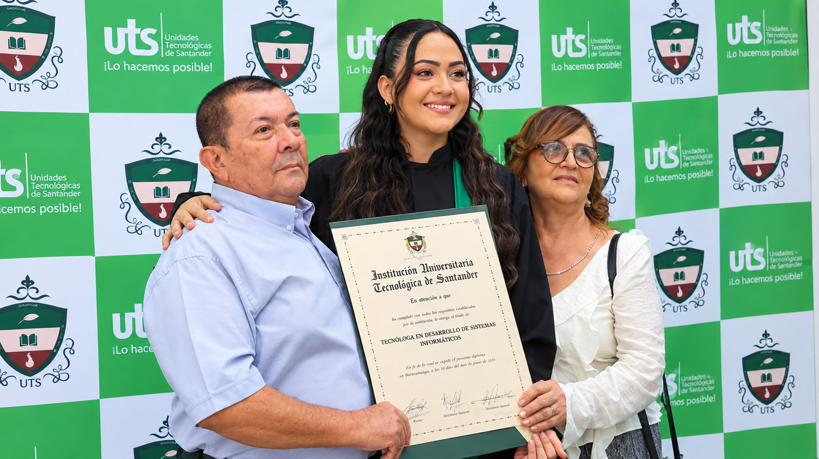 Una graduada sonríe entre sus padres y sostiene su diploma de Tecnóloga en Desarrollo de Sistemas Informáticos, expedido por la Institución Universitaria Tecnológica de Santander.